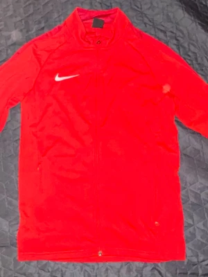 Röd Nike zip-tröja med logga - Säljer en röd zip-tröja från Nike med vit logga på bröstet. Tröjan har ståkrage och hel dragkedja framtill. Perfekt för träning eller chill, med lång ärm och sportig look. Materialet känns mjukt och lätt mot huden.