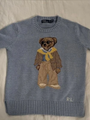Blå stickad tröja Polo Bear Ralph Lauren - Säljer en ljusblå stickad tröja från Polo Ralph Lauren med ikonisk Polo Bear-motiv framtill. Tröjan har ribbade muddar vid ärmslut och nederkant, rund hals och RL-broderi nedtill. Perfekt för dig som gillar klassisk stil med en twist.