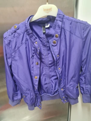 Lila jacka från DIVIDED - Snygg lila bomberjacka från DIVIDED med guldiga knappar och dragkedja. Jackan har ribbade muddar vid ärmslut och nederkant, samt coola axeldetaljer. Perfekt för dig som vill sticka ut med färg och stil.