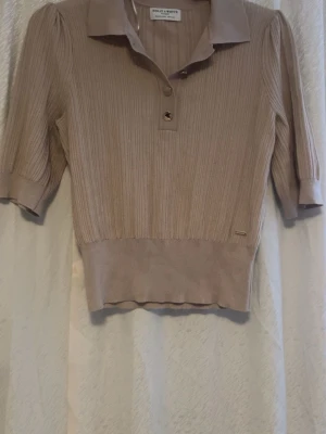 Beige ribbad pikétröja Holly & Whyte - Snygg beige pikétröja från Holly & Whyte med ribbad struktur och korta ärmar. Tröjan har klassisk krage, två knappar framtill och bredare muddar vid ärmslut och nederkant. Perfekt för dig som gillar stilrena och bekväma plagg.