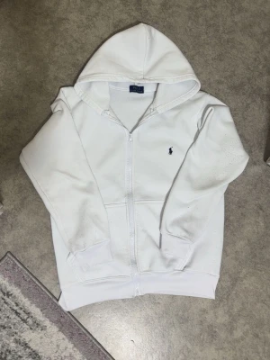 Hoodie från ralph lauren - Jag säljer en vit ralph lauren hooddie. Den är ganska använd men den har ett skönt material och kan kålla dig varm. OBS! Den är inte äkta