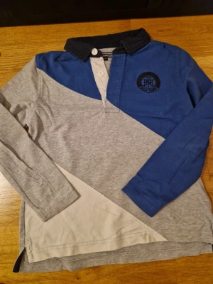 Blå och grå pikétröja Tommy Hilfiger - Snygg pikétröja från Tommy Hilfiger i blått, grått och beige med mörkblå krage och broderad logga på bröstet. Tröjan har långa ärmar, knappar vid halsen och är gjord i mjuk bomull. Perfekt för dig som gillar klassisk stil med en twist.