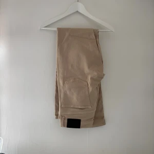 Beiga jlindberg byxor - Snygga beiga cargopants med stora fickor på benen och justerbar kardborre vid benslut. Perfekta för en avslappnad och trendig stil. Tillverkade i ett mjukt bomullsmaterial som är skönt att bära hela dagen.