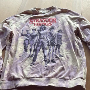 Stranger Things tröja H&M lila/vit - Cool sweatshirt från H&M med Stranger Things-tryck framtill. Tröjan har en batikmönstrad bas i lila och vitt, rund hals och långa ärmar. Perfekt för dig som älskar serien och vill sticka ut med en unik look.