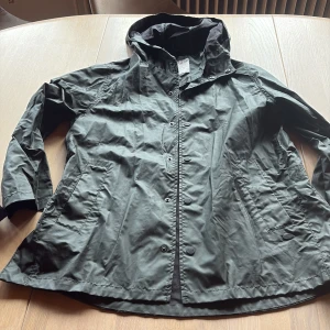 Grön regnjacka från Zara Kids - Snygg grön regnjacka från Zara Kids med huva och praktiska fickor. Jackan har knappar framtill och är tillverkad i ett lätt syntetmaterial som står emot regn. Perfekt för blöta dagar och enkel att slänga på sig när det behövs.