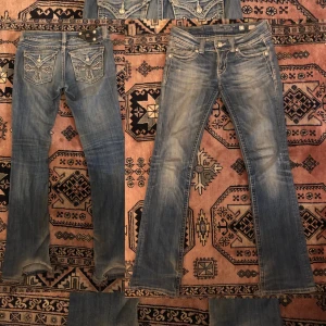 Miss Me bootcut jeans med strass - Snygga blå bootcut jeans från Miss Me med coola strassdetaljer och broderade kors på bakfickorna. Jeansen har låg midja, tydliga vita sömmar, med uttvättning och dekorativa knappar. De är i storlek 27 och jeansen passar storleken bra. Andra bilden är lånad men jag är 168 och de är några centimeter för långa för mig, därav säljer jag de. Som man kan se på bilderna finns det slitningar längst ner som knappt syns. Kontakta mig om du har några frågor.
