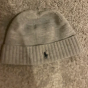 Stickad mössa från Polo Ralph Lauren - Grå stickad mössa från Polo Ralph Lauren med klassisk svart broderad logga framtill. Mössan har bred ribbad kant och är tillverkad i mjukt material som håller dig varm under kyliga dagar. Perfekt accessoar för en clean och stilren look. Saknas lapp på insidan som inte spelar någon roll alls för den delen. Lämna gärna ett eget prisförslag!