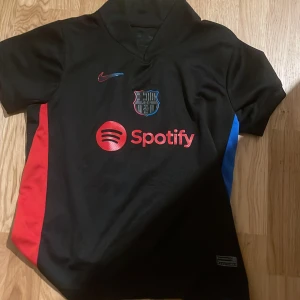 FC Barcelona svart bortatröja Pedri 8 - Säljer en svart FC Barcelona bortatröja från Nike med Pedri 8 på ryggen. Tröjan har röda och blå detaljer på sidorna, klubbmärke och Nike-logga i 3D-effekt samt Spotify som sponsor framtill. Tillverkad i lätt och ventilerande material.