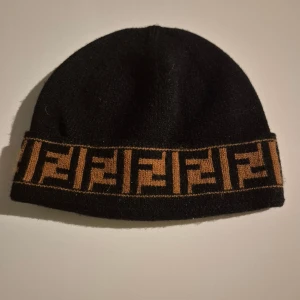 Svart Fendi mössa med logobård - Svart mössa från Fendi med bred brun bård runt kanten, dekorerad med det klassiska FF-logomönstret. Snygg och stilren accessoar som lyfter vilken outfit som helst. Tillverkad i mjukt material som håller dig varm under kyliga dagar.