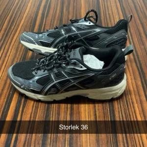 Svarta Asics sneakers storlek 36 - Snygga svarta Asics sneakers med grå och vita detaljer. Skorna har mesh- och syntetpaneler, snörning och en rejäl, vit sula som ger bra komfort. Perfekta för dig som gillar sportig stil och vill ha ett par bekväma skor till vardagen.