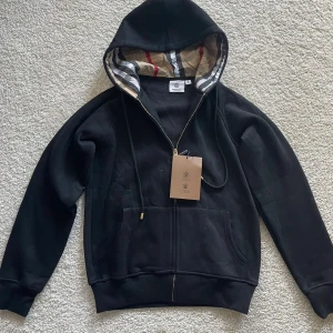 Svart hoodie från Burberry med dragkedja - Svart hoodie från Burberry med klassiskt rutigt foder i huvan, guldfärgade detaljer på snörena och dragkedja framtill. Stor känguruficka och ribbade muddar. Perfekt för dig som vill ha en stilren och lyxig look. Koftan är aldrig använd! Tveka inte att höra av er vid några frågor eller funderinga🤗