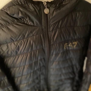 Svart pufferjacka från EA7 Armani - Säljer en svart pufferjacka från EA7 Emporio Armani med guldfärgad logga på bröstet. Jackan har quiltad design, huva och hel dragkedja framtill. Perfekt för dig som vill ha en stilren och modern look med exklusiv känsla.