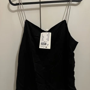 Svart linne med glittrig band - Svart linne från H&M med smala, glittriga axelband. Linnet har en enkel och stilren design med rak passform och ärmlös modell. Perfekt för dig som gillar minimalistisk stil och vill ha något snyggt och enkelt.