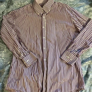 Randig skjorta från Tailor Store - Säljer en snygg skjorta från Tailor Store med lila och vita vertikala ränder. Skjortan har klassisk krage, bröstficka och knäppning framtill. Tillverkad i 100% bomull och har långa ärmar med manschettknappar. Perfekt för dig som gillar stilrena detaljer. storlek står ej men uppskattar på s/m