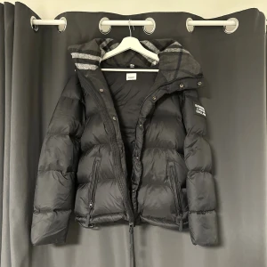 Svart pufferjacka från Burberry - Svart pufferjacka från Burberry med avtagbara ärmar, så den kan användas som väst. Jackan har dragkedja, knappar och två fickor med dragkedja. Insidan av huvan har ett grått rutigt mönster. Patch med Burberry London England på ärmen.