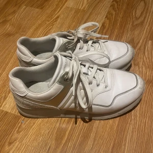 Vita sneakers från Tommy Hilfiger - Snygga vita sneakers från Tommy Hilfiger med silvriga detaljer och logga på sidan. Skorna har snörning, rund tå och platt sula. Tillverkade i skinn och syntet med bekväm textilinsida. Perfekta för dig som gillar stilrena och sportiga vibes.
