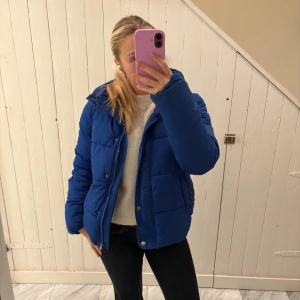 Blå pufferjacka från ONLY - Snygg blå pufferjacka från ONLY i storlek S. Jackan har hög krage, dragkedja och tryckknappar framtill samt två fickor på sidorna. Perfekt för kyligare dagar och ger en cool look till din outfit.