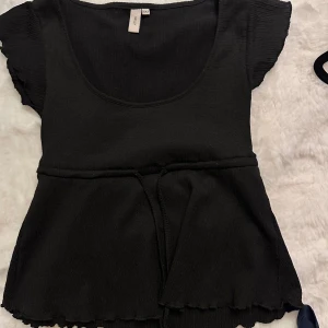 Svart blus med knyt från Nelly - Supersöt svart blus från Nelly med korta volangärmar, markerad midja och knytband. Blusen har en rundad urringning och vågig kant nertill för en extra detalj. Perfekt att styla med jeans eller kjol för en trendig look.