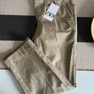 Beige chinos från Zara, strl 36 - Snygga beige chinos från Zara i storlek 36. Klassisk rak passform med fickor fram och bak, bälteshällor och knappstängning. Tillverkade i mjuk bomull som är skön mot huden. Perfekta för en chill och stilren look.