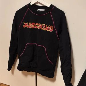 Svart Moschino sweatshirt med rosa detaljer - Cool svart sweatshirt orginal från Moschino med färgglad logga i gult och rött på bröstet. Tröjan har rosa kontrastsömmar och en stor magficka framtill. Långärmad och tillverkad i mjuk bomull, perfekt för dig som gillar streetstyle och designer. Storlek 6 i USA alltså 36-38 EU, man kan använda den om man har S och ibland M. Den är använd ett få tal gånger då den blev för liten för mig snabbt orginal priset var 1300.