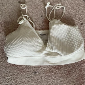 Vit mjuk bhtopp från Kappahl - Supersöt vit mjuk bhtopp från Kappahl med ribbat mönster och smala axelband. Bh-n har v-ringning och en bred resår nedtill för extra komfort. Perfekt till sommarens outfits och enkel att matcha med allt.
