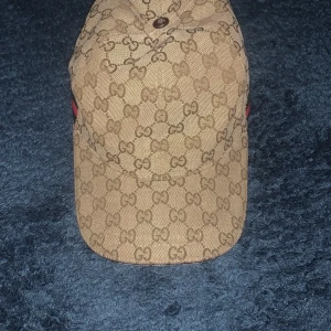 Gucci keps med GG-mönster beige - Snygg keps från Gucci i beige med klassiskt GG-monogram över hela kepsen. Justerbar rem baktill i brunt skinn och diskreta gröna och röda detaljer på sidorna. Tillverkad i bomullscanvas med exklusiv känsla, perfekt för dig som vill sticka ut. Frågor svaras via dm./ mvh