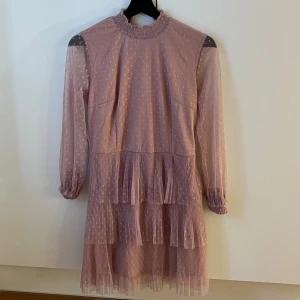 Rosa prickig volangklänning NLY Trend - Supersöt rosa kortklänning från NLY Trend med prickigt mönster, volanger och plisserad nederdel. Hög krage med rynk, långa ärmar med resår vid handlederna och lätt transparent tyg. Perfekt för dig som vill ha en romantisk och trendig look.