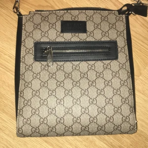  axelväska med GG-mönster - Snygg axelväska  med klassiskt GG-mönster i beige och brunt. Väskan har en svart läderdetalj med dragkedja framtill och en bred axelrem i marinblått och rött. Insidan är fodrad med svart tyg och har en innerficka med dragkedja. Perfekt för dig som gillar designer.