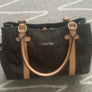Brun handväska från Calvin Klein - Snygg handväska från Calvin Klein i mörkbrunt med beige detaljer och logomönster över hela väskan. Väskan har två rejäla handtag, flera fack och dragkedja baktill. Insidan är fodrad med CK-mönster och har extra fickor för småsaker. Perfekt för dig som gillar stilren design.