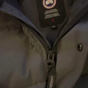  Canada Goose Jacka - Säljer en mörkgrå pufferjacka från Canada Goose med huva och klassisk logga på ärmen. Jackan har dragkedja och knappar framtill, samt stora fickor. Perfekt för kalla dagar och har en riktigt schysst streetstil.
