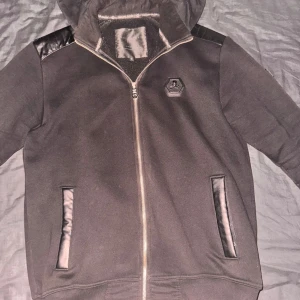 Svart hoodie från Philipp Plein - Svart hoodie från Philipp Plein med huva och dragkedja. Snygga detaljer i skinn på axlar och fickor, samt logotyp på bröstet. Huva med broderad text och ribbade muddar. Perfekt för dig som gillar streetwear med exklusiv känsla.