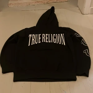 Svart True Religion hoodie med tryck - Svart hoodie från True Religion med stor vit logga på bröstet och tre vita emblem på ena ärmen. Klassisk huva och ribbade muddar. Snygg streetstil och riktigt skön passform. Perfekt för dig som gillar bold prints och vill sticka ut.