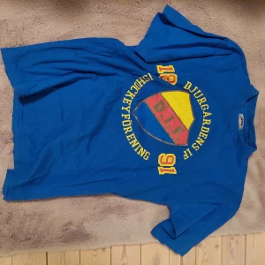 Blå Djurgården t-shirt med tryck - Säljer en blå t-shirt från Djurgårdens IF med stort tryck på bröstet i gult, rött och blått. T-shirten har rund hals och korta ärmar, perfekt för dig som vill visa ditt hockeyintresse. Klassisk supporterstil med DIF-logga och årtal. Från 90 talet. 
