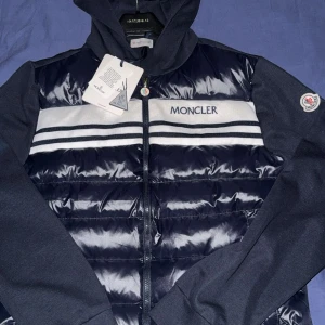 Moncler cardigan med huva - Snygg cardigan från Moncler i mörkblått med vita och blå ränder över bröstet. Jackan har huva, dragkedja framtill och Moncler-logga på ärmen. Materialmix med glansig polyester på kroppen och mjuka bomullsärmar. Perfekt för dig som gillar streetwear.