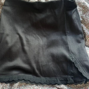 Svart ribbad minikjol med spetskant - Snygg svart kortkjol med ribbad struktur och dekorativ spetskant längst ner. Kjolen har en liten slits framtill som ger extra edge. Perfekt för dig som gillar en stilren men ändå detaljrik look.