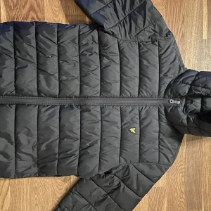 Svart pufferjacka från Lyle & Scott - Svart pufferjacka från Lyle & Scott med gul broderad logga på bröstet. Jackan har huva, hel dragkedja framtill och quiltad design. Materialet är syntet och jackan har en clean, modern look som passar perfekt när det är kallt ute. Har inte den minsta skada (pris går att diskutera vid snabb affär!) Nypris runt 1000kr…