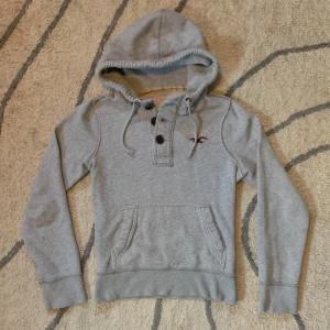 Grå hollister hoodie  - Snygg grå hoodie från Hollister med broderad logga på bröstet och praktisk magficka. Tröjan har huva med snörning och tre knappar vid halsen. Mjuk insida och långa ärmar gör den perfekt för kyliga dagar. Klassisk och enkel stil som funkar till allt. Den är i gott skick men tvättat ett par gånger