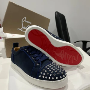 Christian Louboutin sneakers med nitar - Mörkblå sneakers från Christian Louboutin med klassisk röd sula och silverfärgade nitar på tån. Skorna har snörning, rund tå och är tillverkade i mocka med vita gummisulor. Perfekt för dig som vill sticka ut med en lyxig och edgy stil.