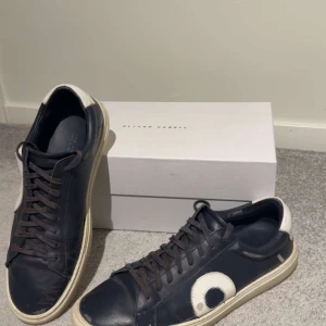 Svarta sneakers från Oliver Cabell - Snygga svarta sneakers från Oliver Cabell med vit detalj på hälen och sidan. Skorna har rund tå, platt sula och snörning. Tillverkade i skinn med en cool kontrast mellan svart och vitt. Perfekta för dig som gillar stilrena och trendiga sneakers. Allt og tillkommer. 