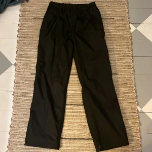 Svarta överdrags byxor  - Svarta överdrags byxor från storm system. Byxorna har två fickor och det finns ett reglerings band i midjan.