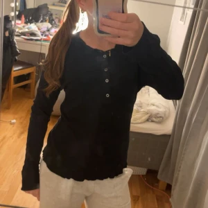 Svart långärmad tröja J.Lindeberg - Svart långärmad tröja från J.Lindeberg i storlek M. Tröjan har rund hals, ribbad struktur och fyra knappar framtill. Passformen är normal och materialet känns mjukt och skönt mot huden. Perfekt för chill dagar eller när du vill ha en enkel stil.