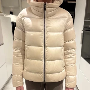 Beige Colmar pufferjacka med huva - Säljer en beige pufferjacka från Colmar Originals med huva och raka, quiltade paneler. Jackan har dragkedja framtill, två sidofickor med dragkedja och Colmar-logga på ärmen. Perfekt för kalla dagar och har en clean, modern look.