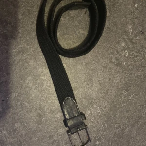 Svart flätat bälte med skinnspänne - Snyggt svart bälte med flätad design och detaljer i svart skinn vid spännet. Bältet har en klassisk metallspänne och passar perfekt till jeans eller chinos. Materialet är mjukt och flexibelt, vilket gör det bekvämt att bära hela dagen.