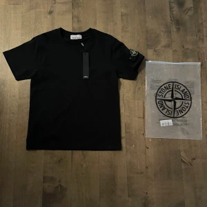 Svart Stone Island t-shirt med patch - Svart t-shirt från Stone Island med klassisk rund hals och korta ärmar. På vänster ärm sitter den ikoniska Stone Island-patchen med broderad logga i gult och grönt, fäst med två knappar. Materialet är mjuk bomull och passformen är normal. Perfekt för dig som gillar streetwear.