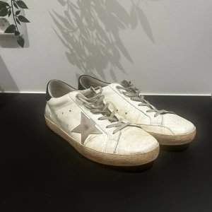 Golden Goose vita sneakers med stjärna - Golden Goose sneakers i vitt skinn med grå mockadetaljer och grå snörning. Sidan pryds av en ikonisk stjärna i grått och texten GGDB/STAR. Baksidan har svart hälkappa med logga. Snygg, vintageinspirerad look och platt sula.