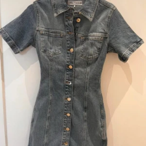 Jeansklänning med knappar från Zara - Snygg jeansklänning från Zara med korta ärmar, krage och knappar hela vägen framtill. Klänningen har två bröstfickor och markerad midja för en slim passform. Perfekt för dig som gillar en cool och avslappnad stil.