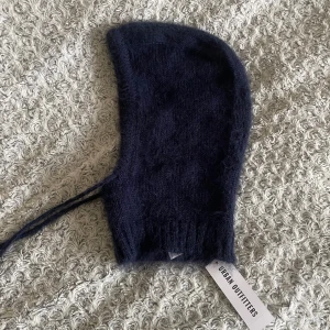 Mörkblå balaclava från Urban Outfitters - Mysig och fluffig mörkblå balaclava från Urban Outfitters. Stickad modell med ribbad kant och långa knytband framtill. Perfekt för kalla dagar och ger en trendig look. Materialet är mjukt och värmande, passar dig som vill ha både stil och komfort.