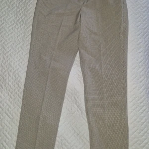 Högmidjade rutiga kostymbyxor - Helt nya högmidjade kostymbyxor i beige och lite rutigt mönster. Byxorna har klassisk dragkedja, knäppning och hyska fram. Snygg passform med raka ben och diskret rutmönster som ger en stilren look.