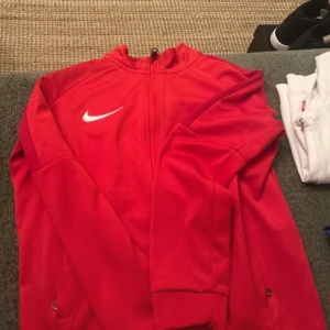 Röd Nike Dri-Fit vindjacka XS - Nike Dri-Fit vindjacka i rött med vit swoosh på bröstet. Jackan har hel dragkedja framtill, sidofickor med dragkedja och sportig design. Tillverkad i lätt polyester som andas, perfekt för träning och aktiva dagar.