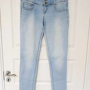 Jeans / legging med bred linning - Ett par ljusblå jeans / legging i tunt och mjukt material (79 % bomull, 19 % polyester, 2 % elastan). Bred linning med tre knappar. Kort dragkedja i gylfen. Slitning i grenen (bild 4). Plagget har naturligtvis tvättats. Se bilder för skick och passform. Storlek L/XL enligt lappen. Midjemått cirka 39 cm.